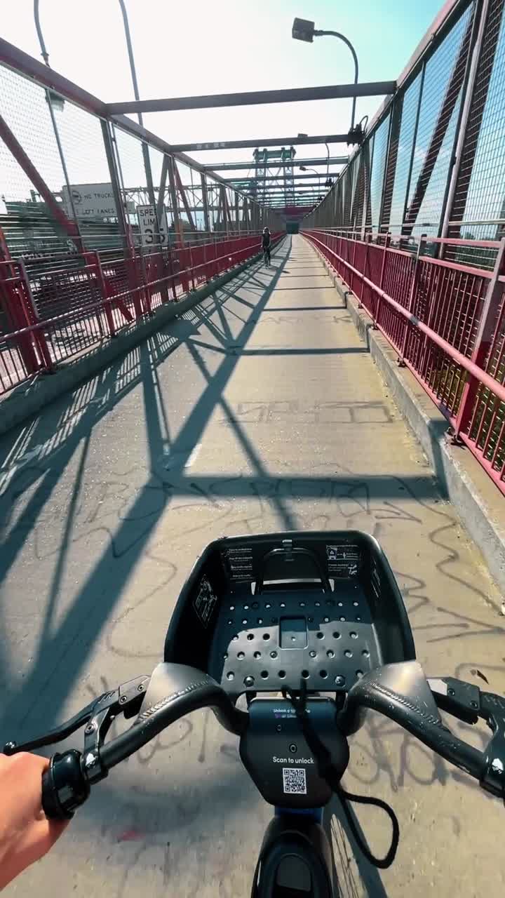 New York, USA, 2 marzo 2026: Punto di vista di una persona che va in bicicletta su una pista ciclabile di un ponte. Scatto POV dal manubrio di una bici elettrica che attraversa un ponte con strutture metalliche e ombre sul selciato in città