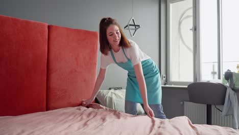 a brunette cleaning lady girl in a white t-shirt and blue apron makes the bed with a pink blanket and gray pillows while cleaning the bedroom when the cleaning company was called