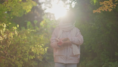 woman walking along autumn garden path flipping pages of book held in hand, warm sunlight filtering through golden foliage, scarf and jacket framing scene as fallen leaves rustle under soft breeze