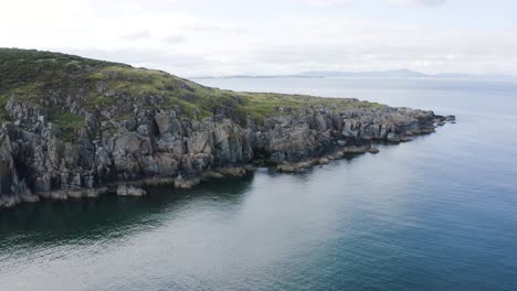 Flying-away-from-the-rocky-coastline-of-Clogherhead-with-the-calm-blue-ocean-on-a-sunny-day,-Ireland