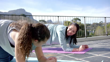 Happy-biracial-mother-and-daughter-practising-yoga-on-terrace-in-sunny-day,-slow-motion