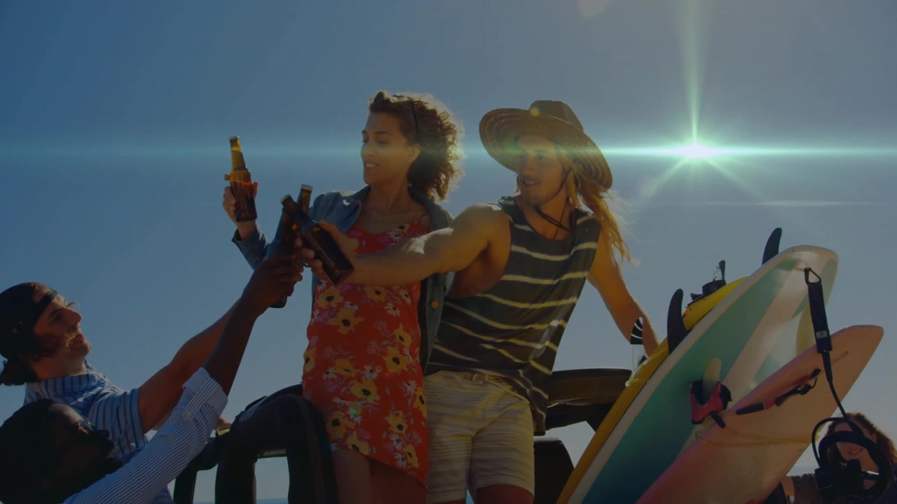 Pubblicità di viaggio che mostra 5 amici che brindano in jeep con tavole da surf in riva al mare, donna che alza una bottiglia accendendo un razzo.