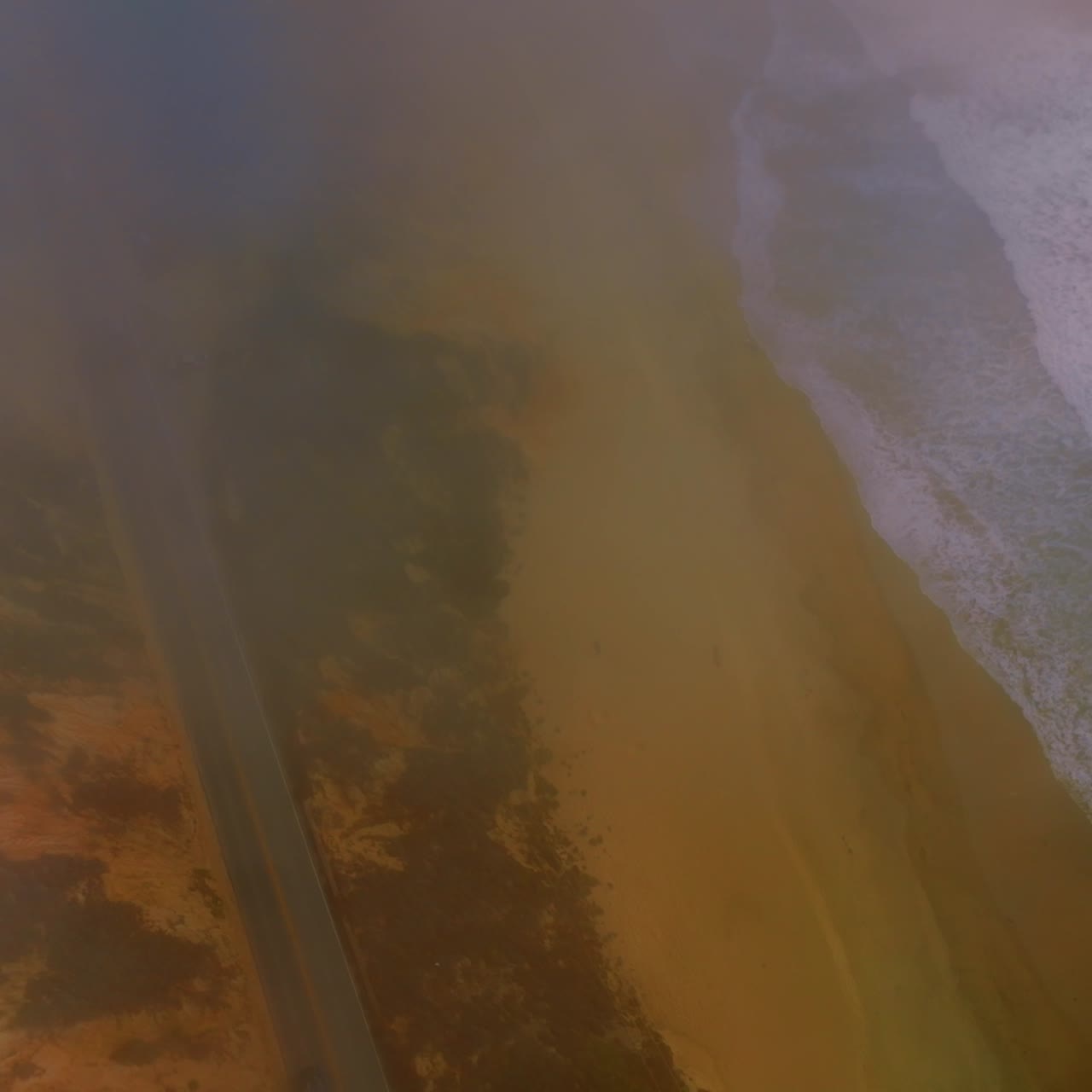 Foamy waves coming to the sandy shore. Drone descending through the fog over the road going by the beach