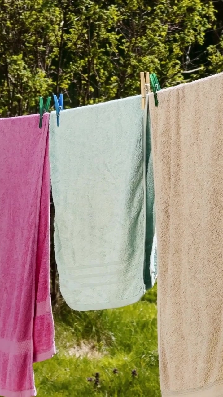 Vertical close-up of beige towels drying on a backyard clothesline with colorful pegs, striped fabric and soft spring garden branches in sunlight.