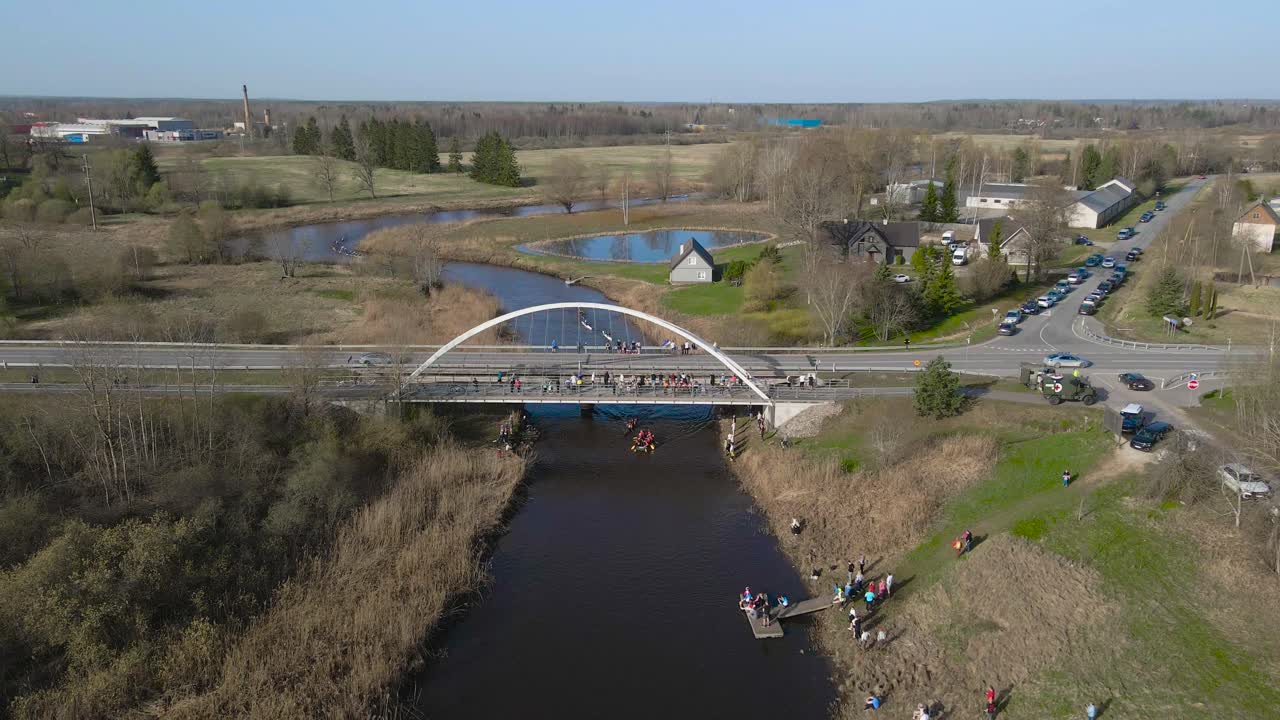 Aerial drone footage showing people paddling and rowing in kayaks, rafts and paddleboards during a sunny day from under a bridge where cars drive at Võhandu marathon. Shoreline is grassy, sky is blue.