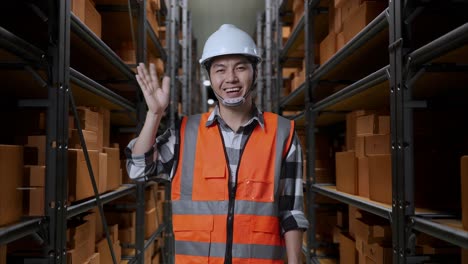 asian male engineer with safety helmet standing in the warehouse with shelves full of delivery goods. smiling to camera and waving hand saying good bye in the storage