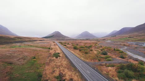 Aérea:-Revelación-De-Un-SUV-Plateado-Que-Viaja-A-Lo-Largo-De-La-Carretera-De-Circunvalación-De-Islandia,-Que-Es-Una-Carretera-Panorámica-Que-Atraviesa-Una-Pintoresca-Y-Remota-Zona-De-Fiordos-Que-Conduce-A-Picos-Gemelos-Con-Niebla-Y-Neblina-En-La-Distancia.