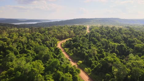 SUV-drives-along-dusty-dirt-road-on-sunny-day-in-tropical-forest,-drone-high-angle-overview