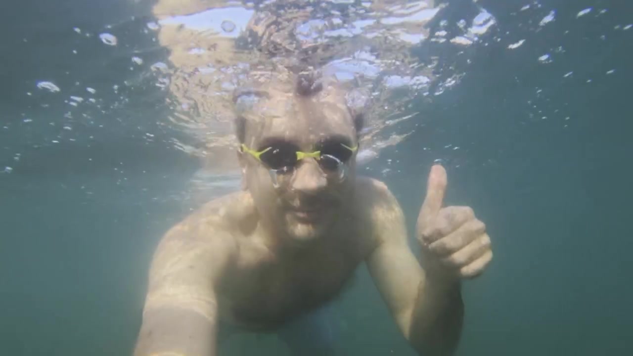 Hombre con gafas nadando bajo el agua, haciendo un gesto de aprobación con el pulgar hacia arriba, creando burbujas en agua azul. Mostrando éxito y positividad.