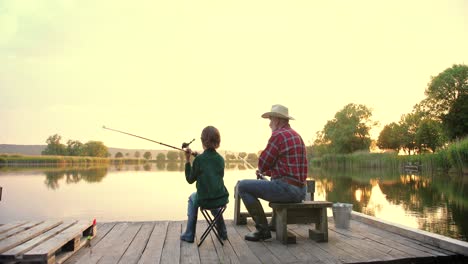 Vista-Trasera-De-Un-Adolescente-Sentado-Con-Su-Abuelo-En-El-Muelle-Del-Lago,-Hablando-Y-Pescando-Juntos