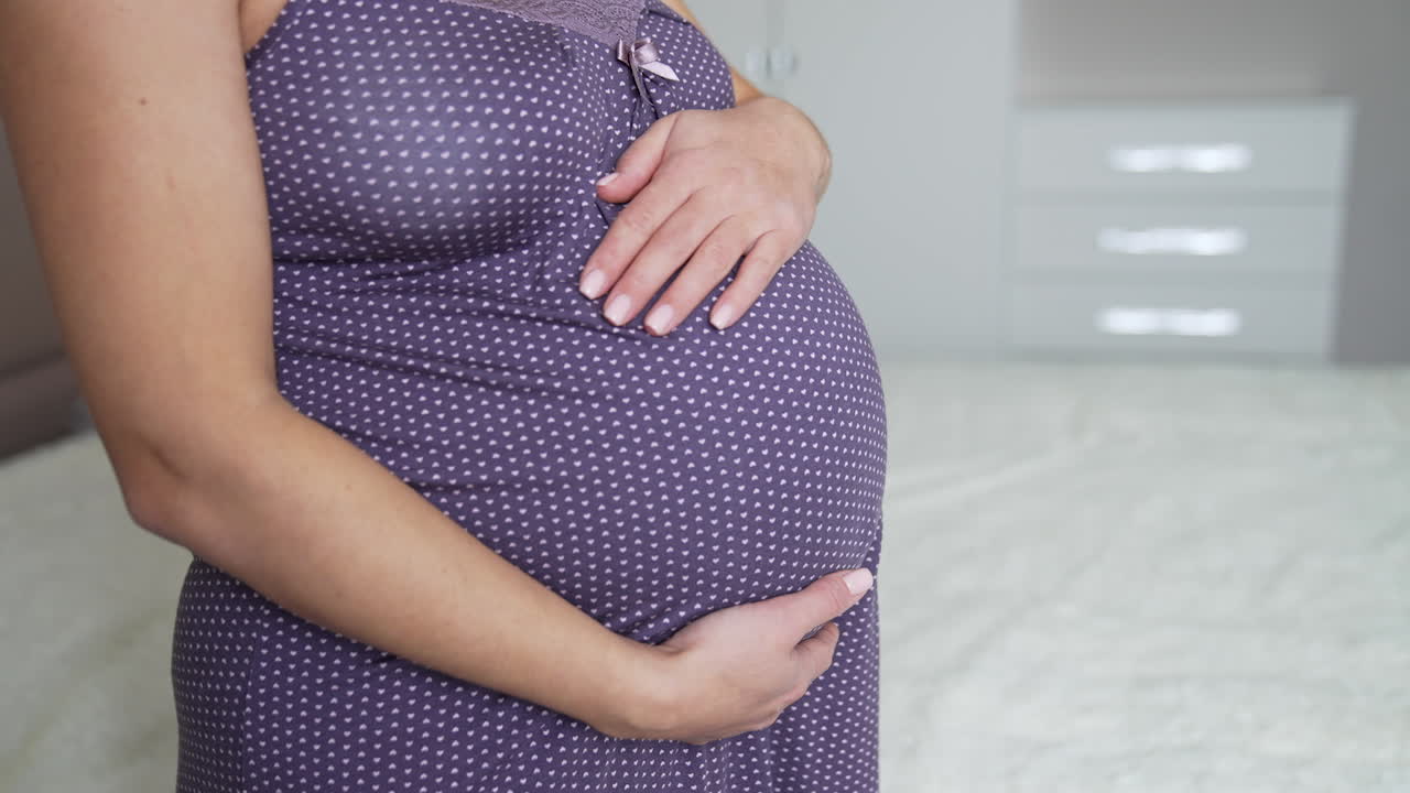 Pregnant woman stroking her big belly with affection. Woman in a night dress stands near the bed in her bedroom. Gentle hands touching big belly lovingly. Close up.