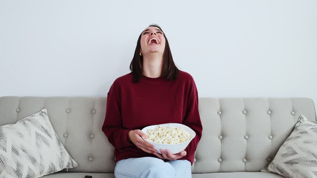 Giovane donna che ride e mangia popcorn sul divano.