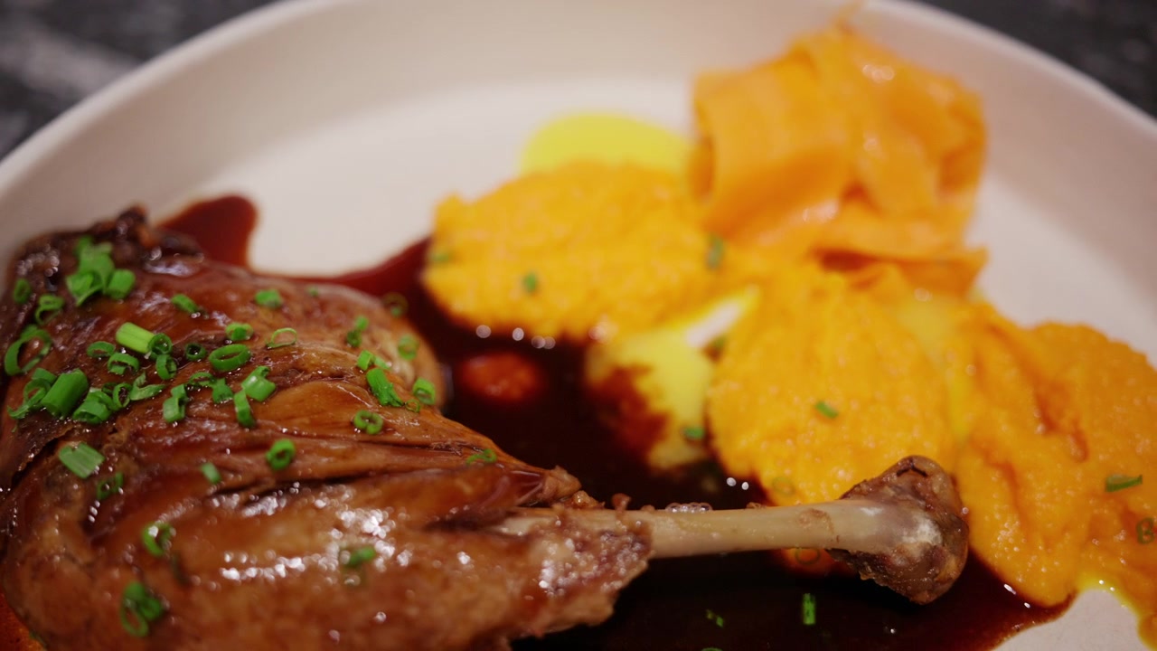 Close-up de uma coxa de pato assada gourmet servida com purê de cenoura sedoso e molho marrom rico em um ambiente de restaurante moderno.