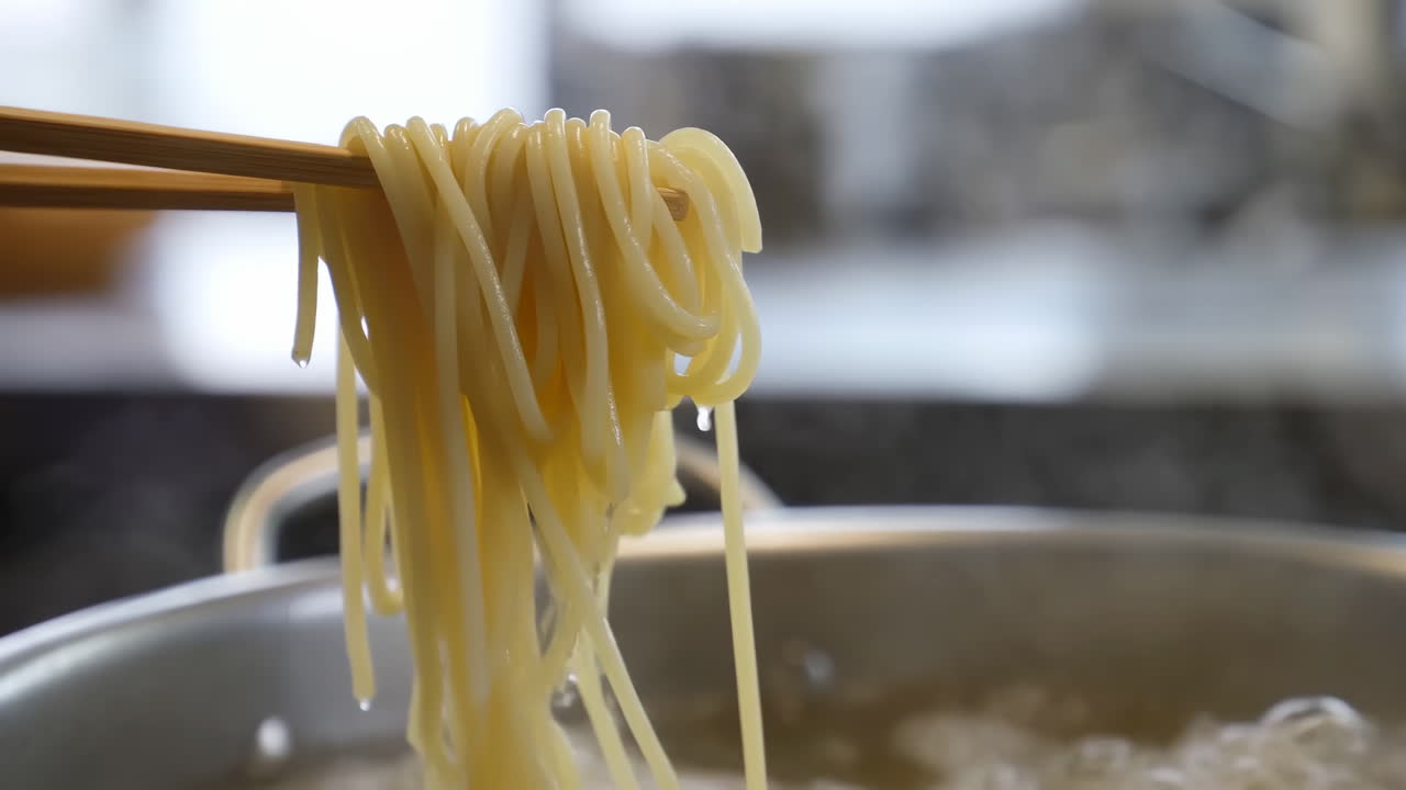 Bacchette che sollevano noodles sopra una pentola bollente, vapore che sale, fili che gocciolano nell'acqua che ribolle, cucina urbana.