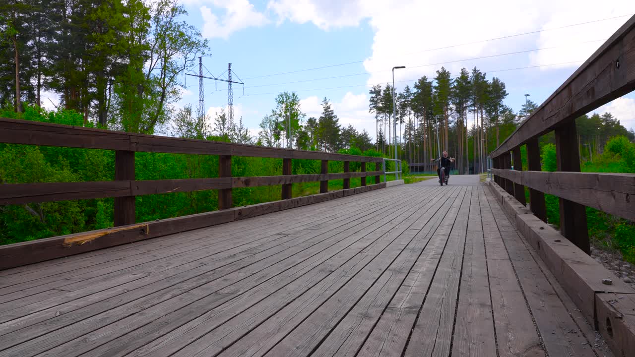 uomo tatuato con abiti neri che cavalca su un ponte di legno con la sua bicicletta in stile elicottero che ha grandi pneumatici whitewall durante l'estate in rallentatore con alberi sullo sfondo in 4k