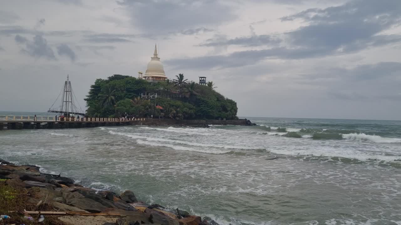 Sri Lanka, Matara’da küçük bir adada yer alan Parewi Duwa Tapınağı’nın beyaz stupasının bulutlu muson gökyüzüne karşı görüntüsü. Çekimde Hint Okyanusu kıyıya vuruyor ve bağlantı...