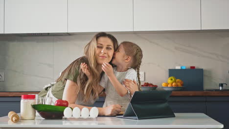 niña pequeña besos y susurros a mamá en la mesa en la cocina