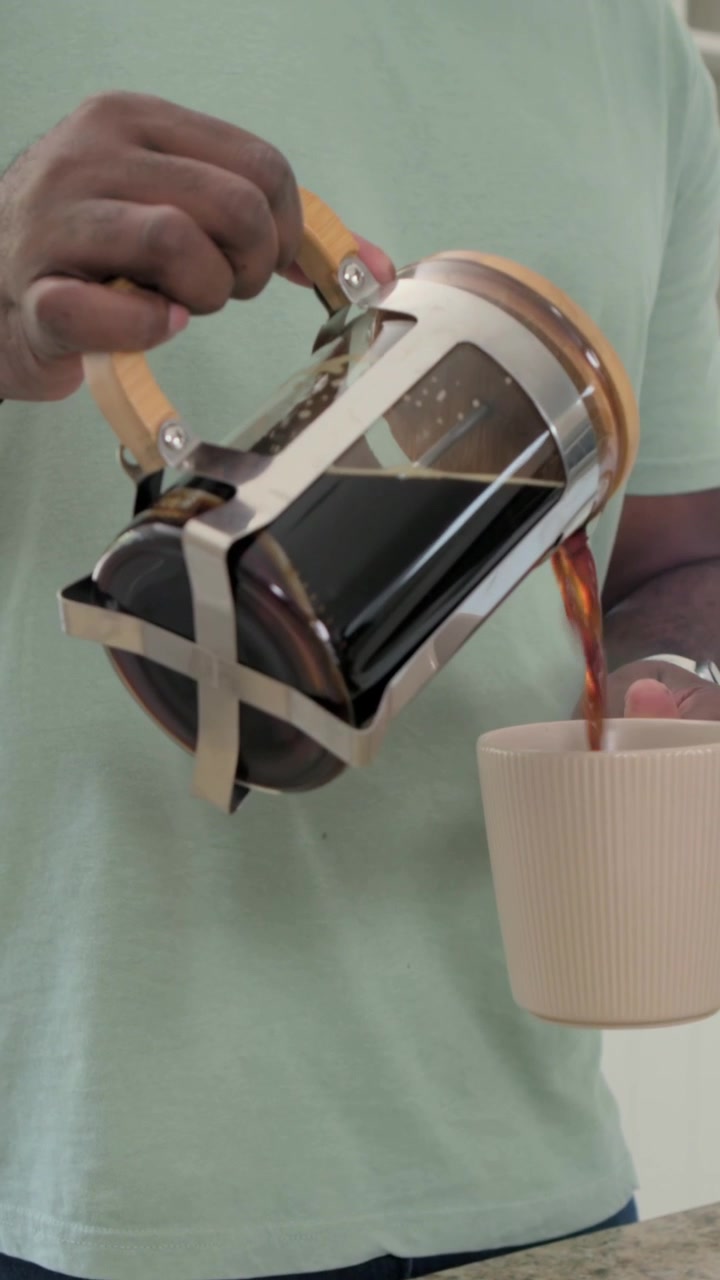 Video verticale: Inclinando la caffettiera, uomo con maglietta verde che versa caffè in una tazza in cucina portandola in una stanza.