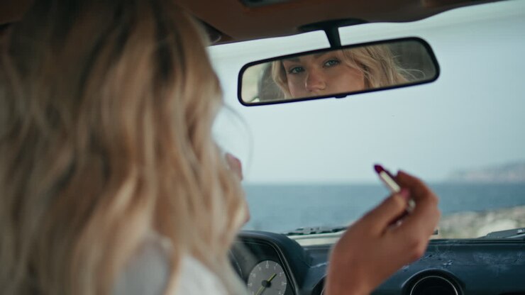 Donna seducente che si guarda nello specchietto dell'auto controllando il trucco, primo piano. Ragazza che si sistema l'acconciatura.