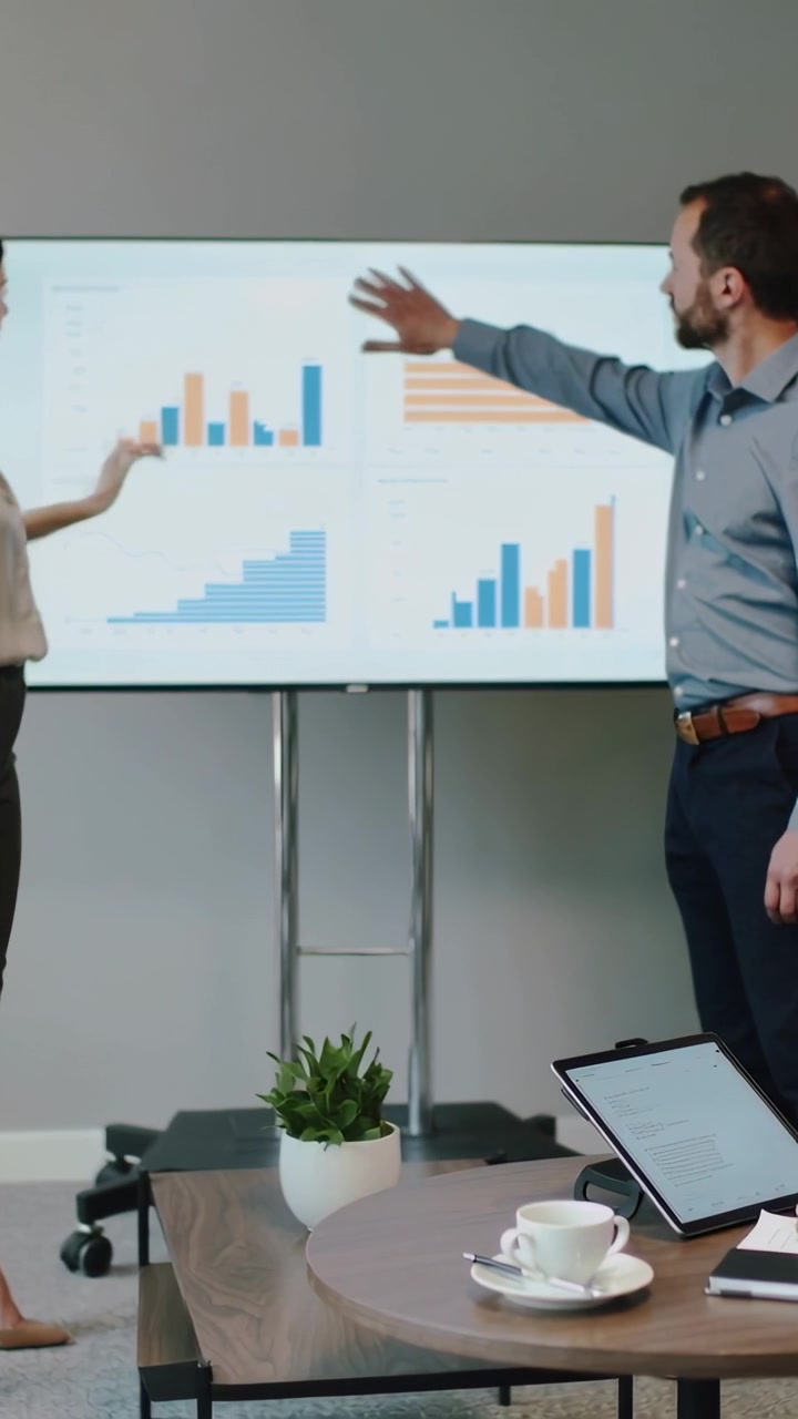 Video verticale: Coppia in abbigliamento da ufficio che fa avanzare una diapositiva, indicando grafici sullo schermo e spiegando i dati