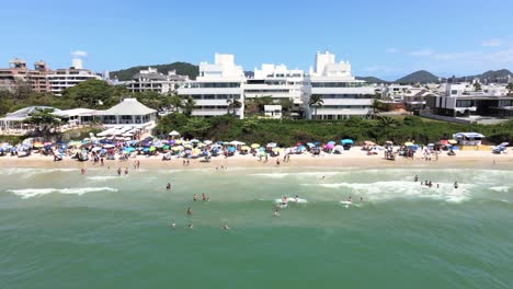 Escena-Aérea-De-Drones-De-Playa-Turística-De-Lujo-En-Florianópolis-Brasil-Con-Mucha-Gente-Y-Construcciones-En-La-Playa-Y-Edificios-Frente-Al-Mar-Con-Hermoso-Océano-Azul-Jurere-Internacional