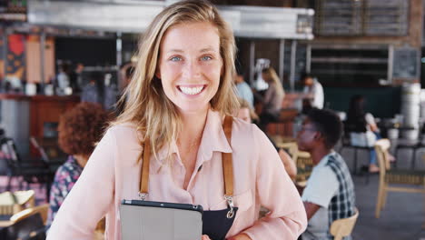 porträt einer kellnerin, die ein digitales tablet in einem belebten barrestaurant hält und zur kamera lächelt