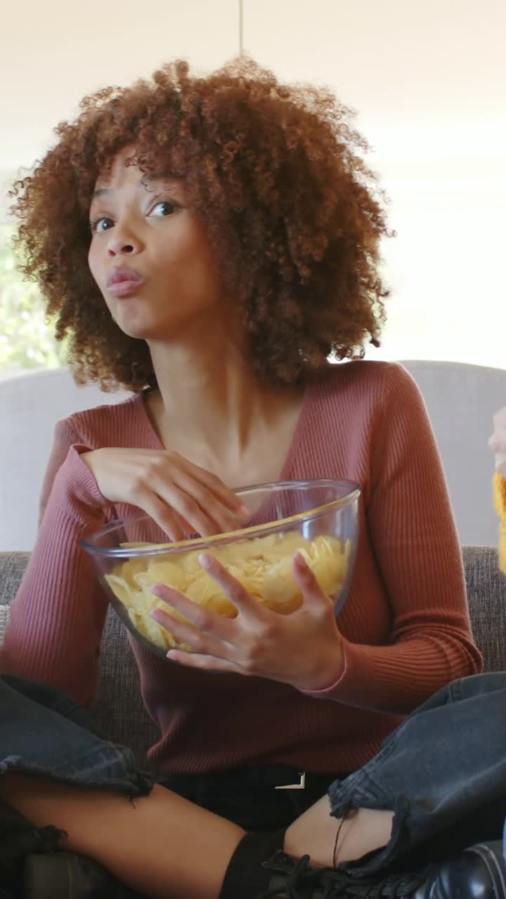 Vídeo vertical: Mulher de top bordô comendo e segurando uma tigela em casa, enquanto um amigo joga videogame com controle.