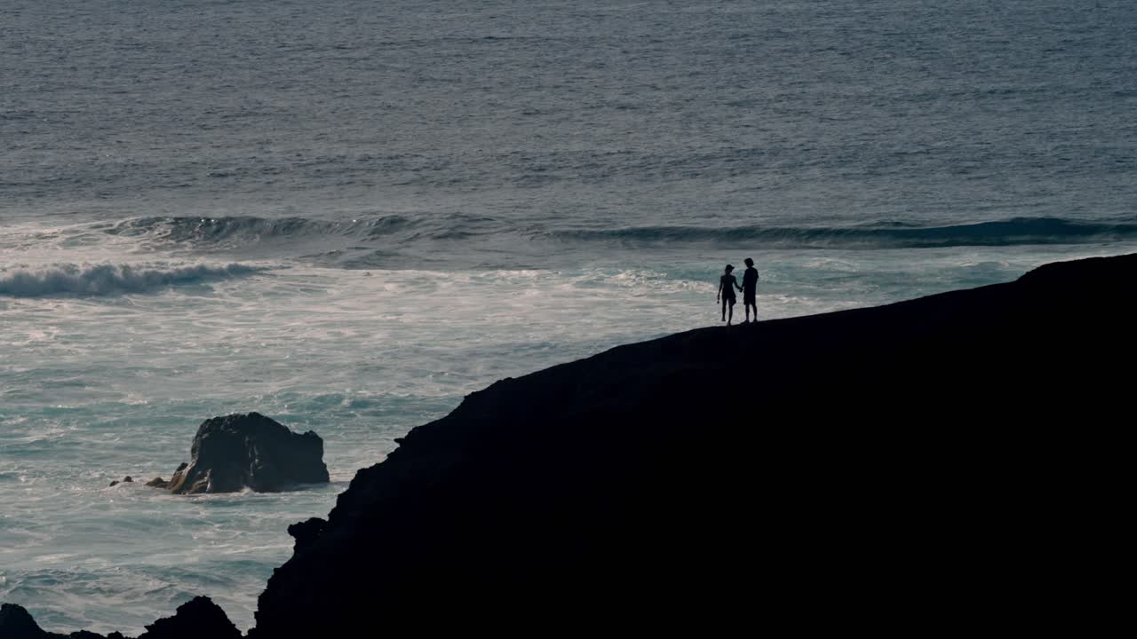 A young couple walks hand in hand along a rugged volcanic cliff overlooking the Atlantic Ocean in Lanzarote, Canary Islands, Spain.