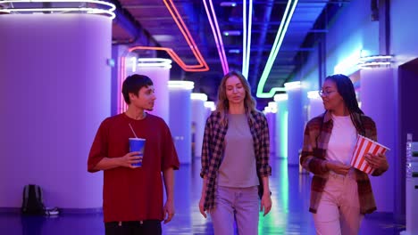 group of three friends walking by cinema hall with neon interior with drink and popcorn bucket