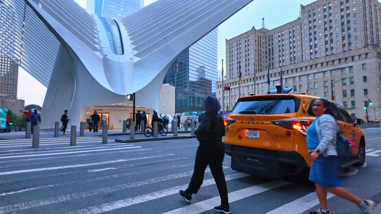 New York, USA, 5 June 2026 : Pedestrians crossing at Oculus entrance with yellow taxi, lower Manhattan. Crosswalk, orange cab, white transit wing overhead and stone office buildings at dusk.