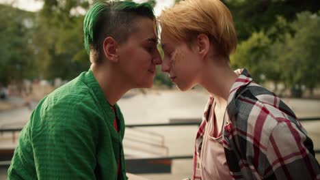 Two-lesbian-girls,-a-girl-with-a-green-haircut-and-green-hair-in-a-green-shirt-and-a-girl-with-yellow-hair-in-a-plaid-shirt,-lean-their-faces-against-each-other-and-rub-their-noses-during-their-date-at-a-skate-park-in-the-summer