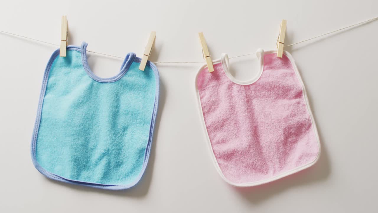 Video of close up of blue and pink bibs with pegs hanging on white background