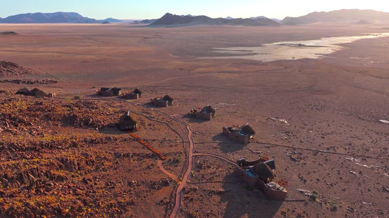 Vista drone di un lodge remoto nel deserto con chalet dal tetto di paglia sparsi nelle pianure rosse della Namibia alla luce dorata. Un rifugio isolato situato nel cuore della natura selvaggia del deserto.