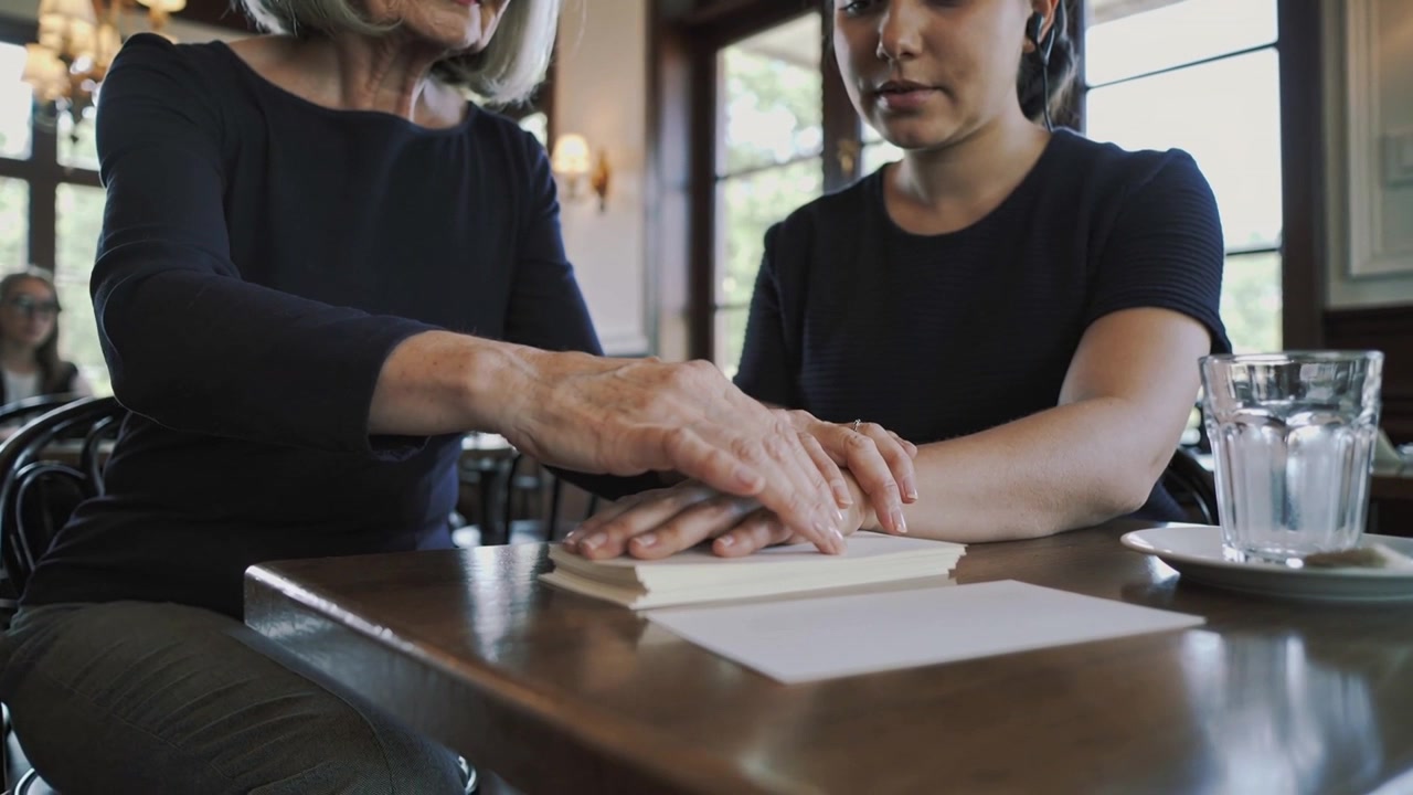 Una donna anziana posa le mani su quelle di un'altra donna a un tavolo di un caffè, confortandola sopra un libro chiuso.