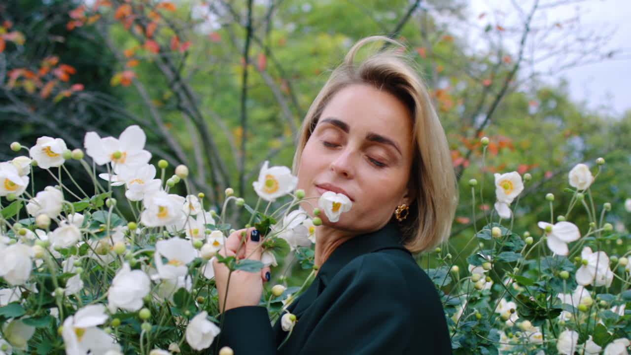 Kvinne med lukkede øyne lukter hvite blomster utendørs. Nærbilde av en blond kvinne med lukkede øyne som lener seg mot hvite anemoner i en hage i New York City