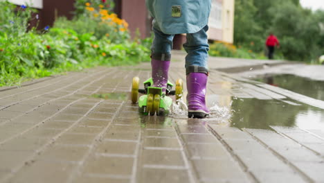 child rides kick scooter on rainy day playful kid wearing rubber boots enjoys scooter riding on wet