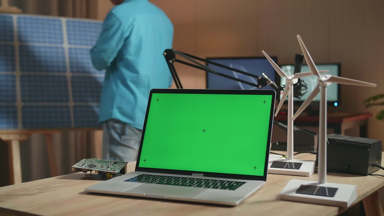 portátil de pantalla verde con placa de circuito y turbina eólica en la mesa mientras un hombre entra en la oficina para ver la célula solar