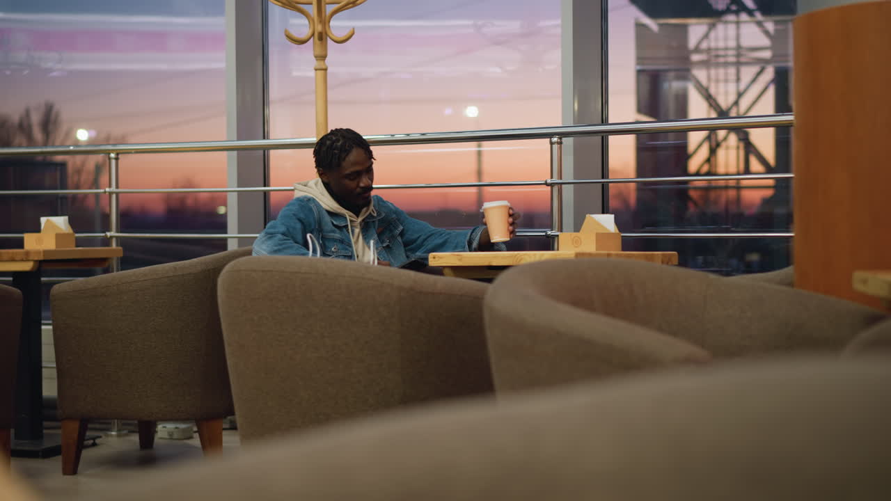 Young man in denim jacket and layered hoodie drinks from paper coffee cup while sitting quietly in cozy lounge near window, surrounded by cushioned chairs, as warm sunset light glows