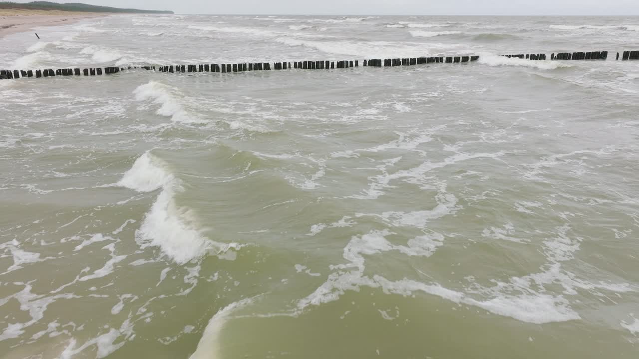 Establishing aerial view of Baltic sea coast on a overcast day, old wooden pier, white sand beach, large storm waves crushing against the coast, climate changes, wide drone shot moving forward low
