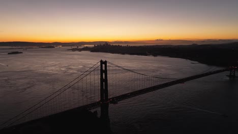 silhouette of golden gate bridge over calm san francisco bay with sunrise horizon and oakland in the distance