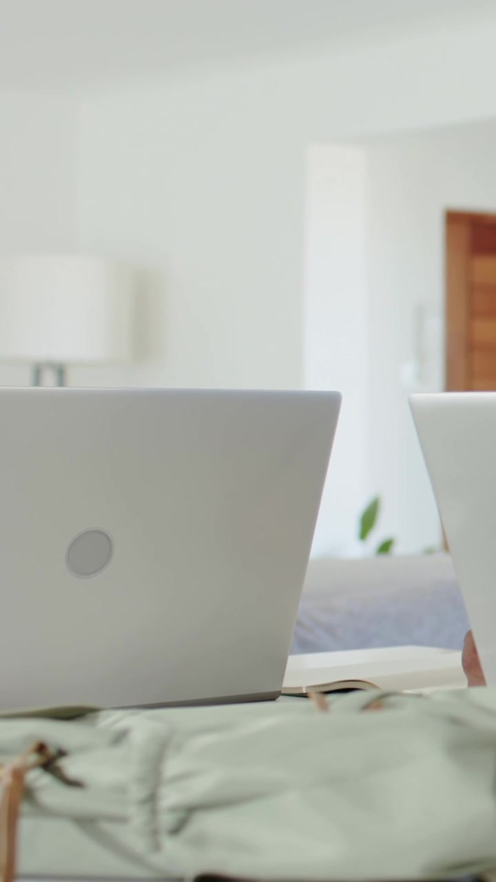 Vertical video: Panning left silver laptop by bed, hand reaching right laptop to use mint backpack