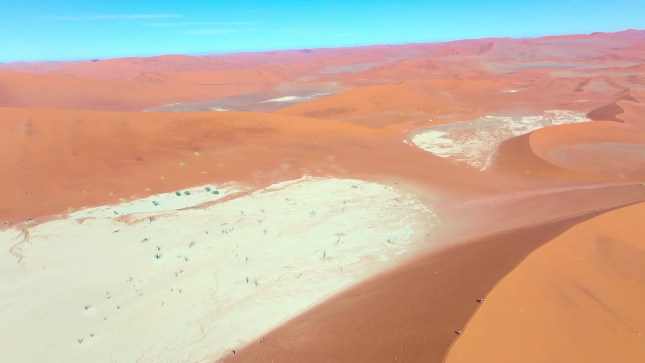 Vista de drone de pessoas atravessando a planície de argila branca de Deadvlei entre as árvores mortas e dunas vermelhas. Pequenos visitantes são minimizados pelas imensas dunas vermelhas e pela planície branca pontilhada com árvores mortas