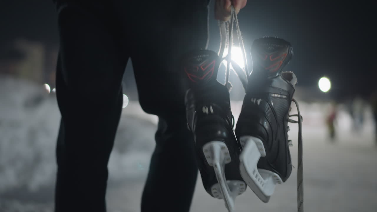 close up of skater walking with pair of ice boots hanging by laces in hand, snowy ground underfoot and soft evening lights in background