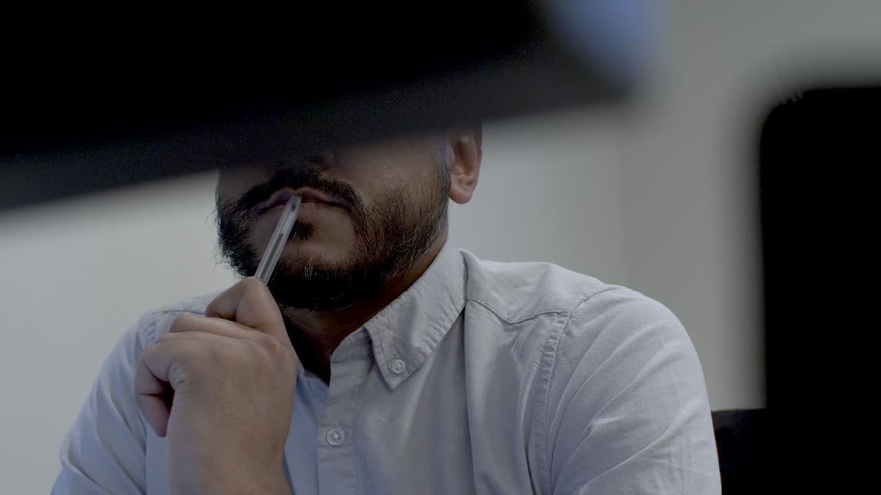 Abstract View Of Bored Employee Tapping Pen On Lips At Desk, Viewed behind two computer monitors. Low Angle, Static, Shot