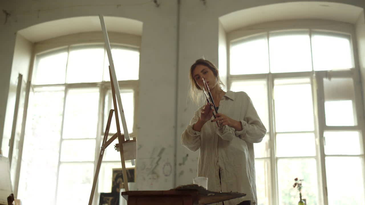 Donna di talento che tiene i pennelli all'interno. Pittrice creativa in piedi vicino al cavalletto.
