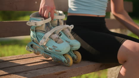 close up of young lady dropping cyan roller skates on wooden bench as she sits beside it, abdomen slightly exposed while relaxed pose and blurred greenery background