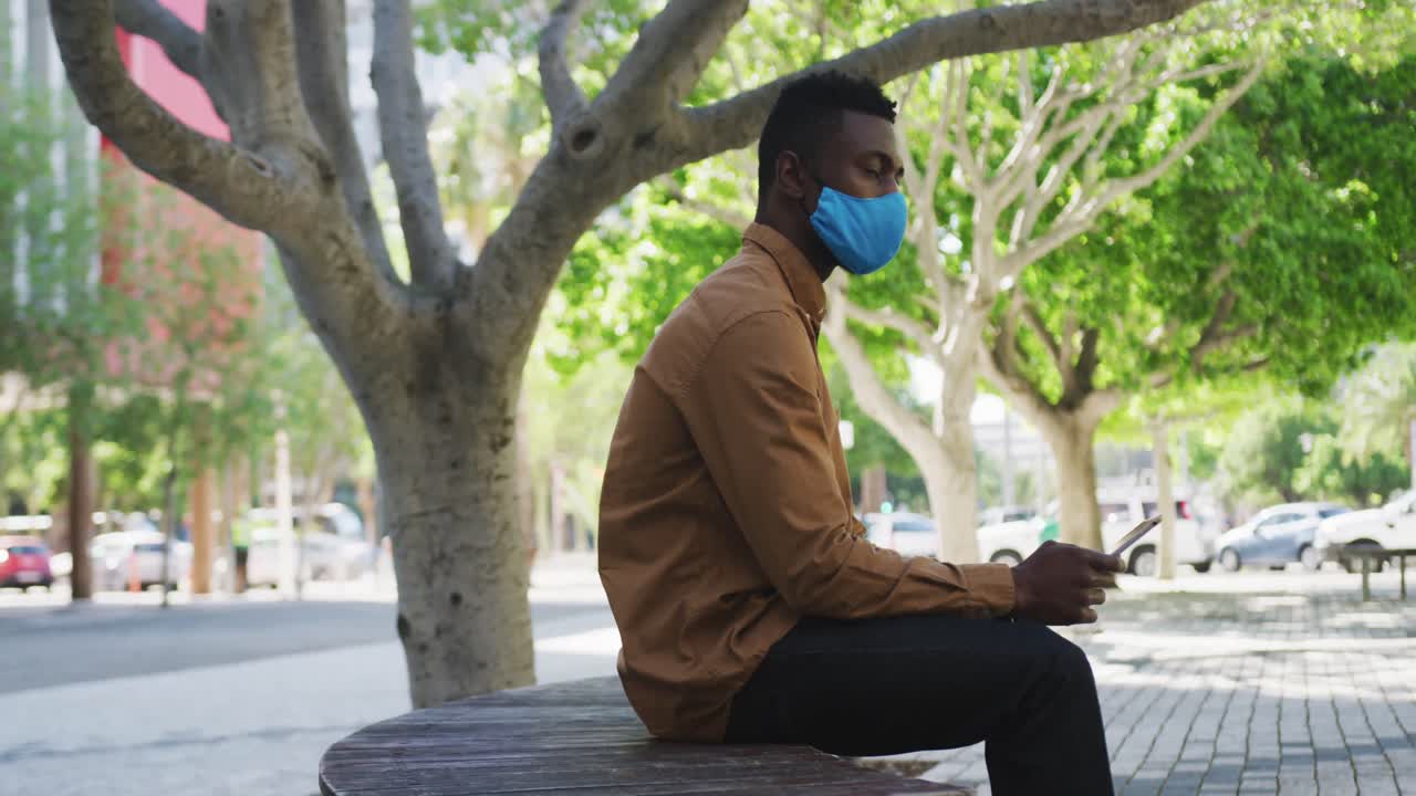 African american businessman wearing face mask using tablet sitting on bench