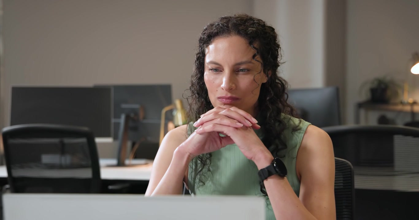Donna afroamericana con un top verde alla scrivania, concentrata sul laptop, si strofina il naso, digita, con uno smartwatch.