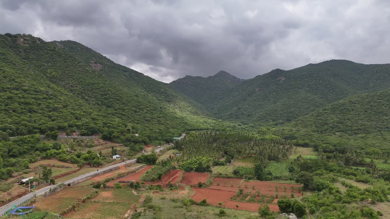 Dramatic aerial view of a mountain range under a heavy, overcast sky. The scene features dense green forests meeting terraced farmland and a road on the valley floor
