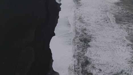 Aerial-top-view-of-ocean-waves-crashing-on-Iceland-Sólheimasandur-black-sand-beach,-on-a-moody-evening